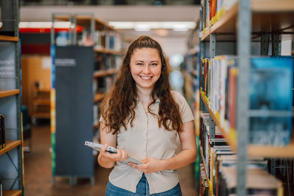Eine junge Frau steht mit einem Tablet in der Hand zwischen Bücherregalen