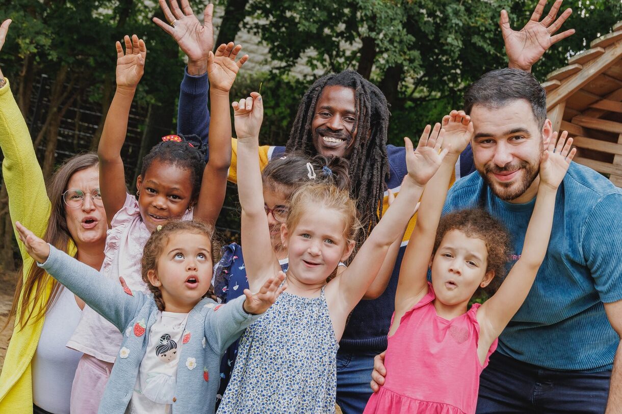 Fünf Kinder stehen mit Erziehungspersonal auf einem Spielplatz, strecken die Hände in die Höhe und lachen