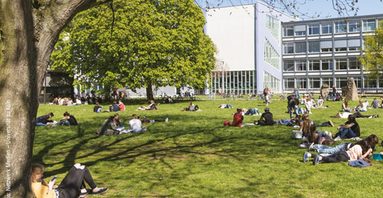 Campus Uni Köln, junge Menschen sitzen auf Wiese mit Bäumen, Gebäude im Hi