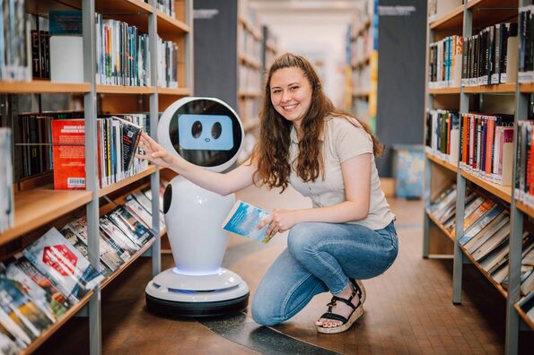 Eine Frau hockt in der Bibliothek neben einem Roboter