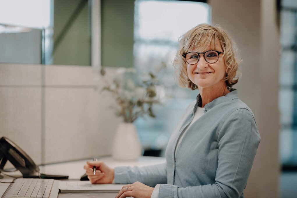 Eine weibliche Person sitzt am Schreibtisch, hält einen Stift in der Hand und lächelt