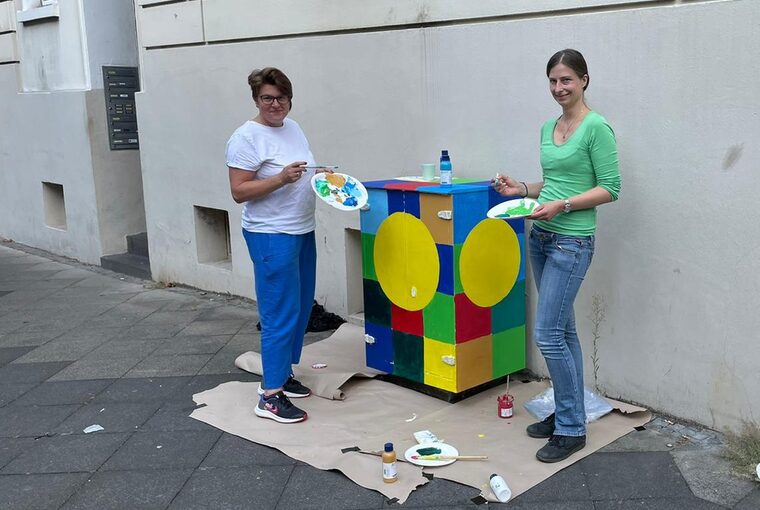 Zwei weibliche Personen bemalen einen Kasten vor einer Hauswand