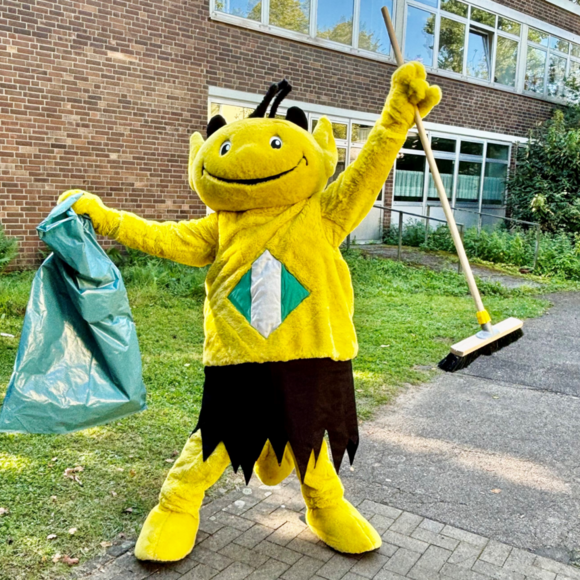 Das Putzteufelchen von Leverkusen hält einen Müllsack und einen Besen in der Hand