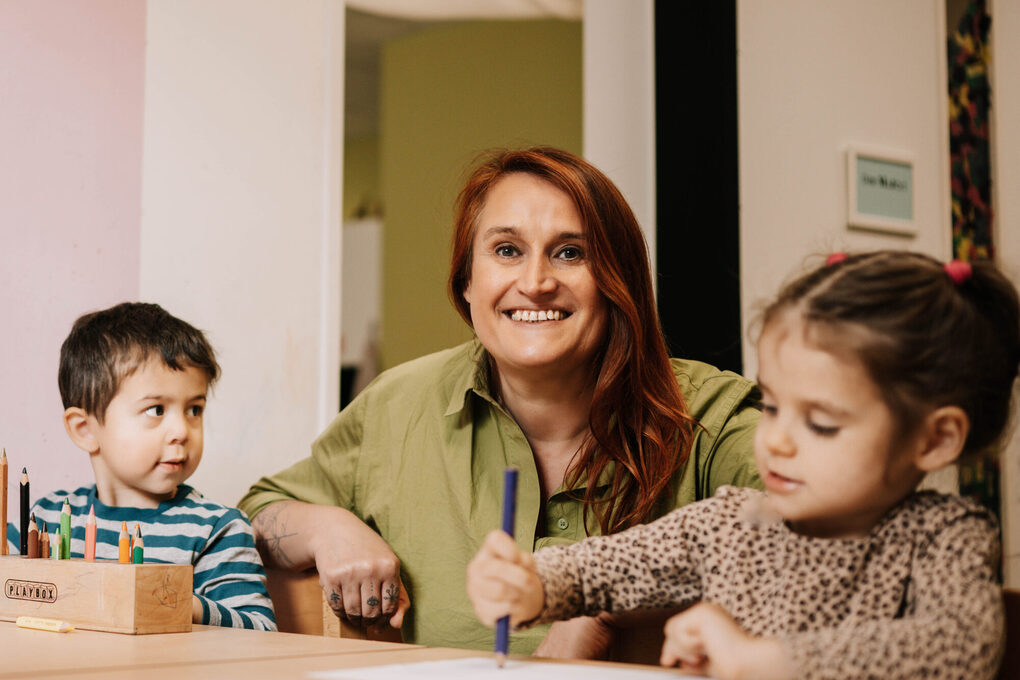 Eine weibliche Person sitzt mit zwei Kindern, die malen, am Tisch