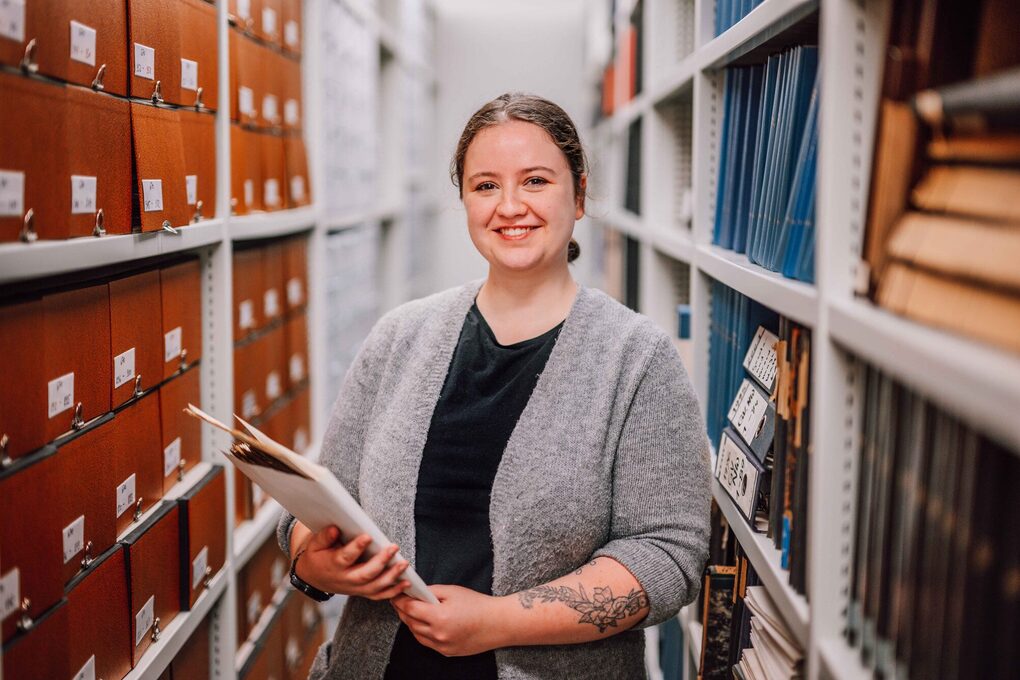 Eine junge Frau steht mit einer Arbeitsmappe zwischen Bücherregalen