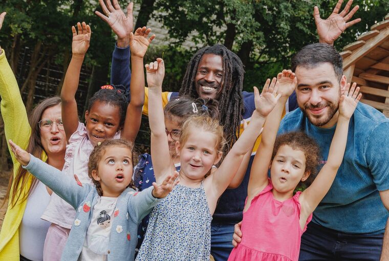 Fünf Kinder stehen mit Erziehungspersonal auf einem Spielplatz, strecken die Hände in die Höhe und lachen